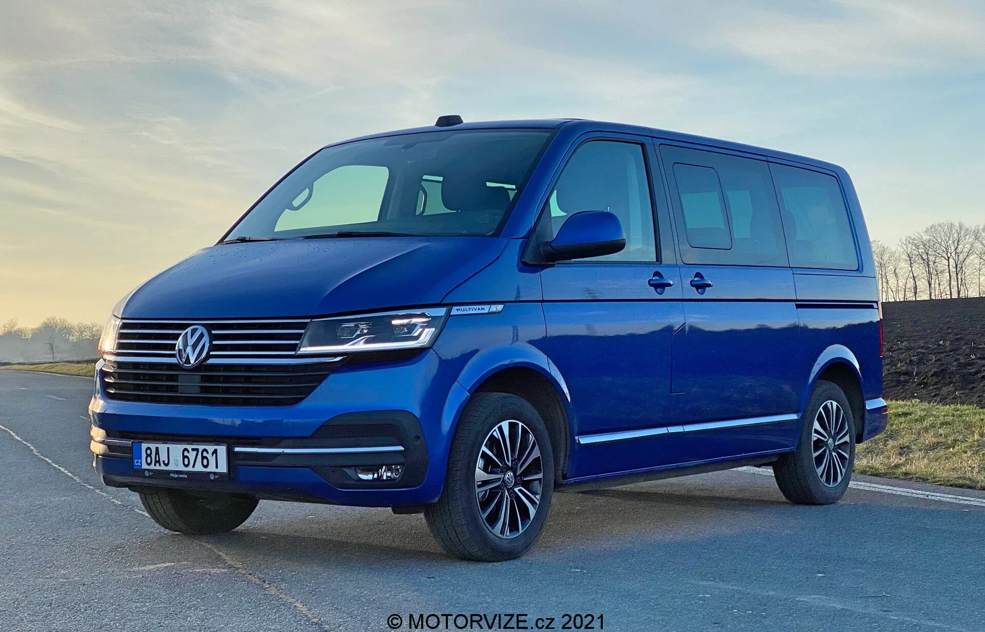 Imagen de una furgoneta azul Volkswagen Transporter T6.1 Multivan de 2019, tomada desde un ángulo bajo para acentuar su estatura y tamaño. La vista de tres cuartos frontal muestra el diseño frontal y lateral del vehículo, incluida la rejilla distintiva de Volkswagen con líneas horizontales, el emblema central, faros modernos que probablemente incluyen tecnología LED o Xenón, y un parachoques delantero elegante con una amplia entrada de aire. Los espejos laterales aerodinámicos con señales de giro integradas, el amplio espacio de las ventanas con una puerta lateral deslizante, llantas de aleación con neumáticos de perfil bajo y una iluminación suave destacan la practicidad y el atractivo estético de la furgoneta. La marca de agua 'MOTORVIZE.cz 2021' sugiere que la imagen es de 2021.