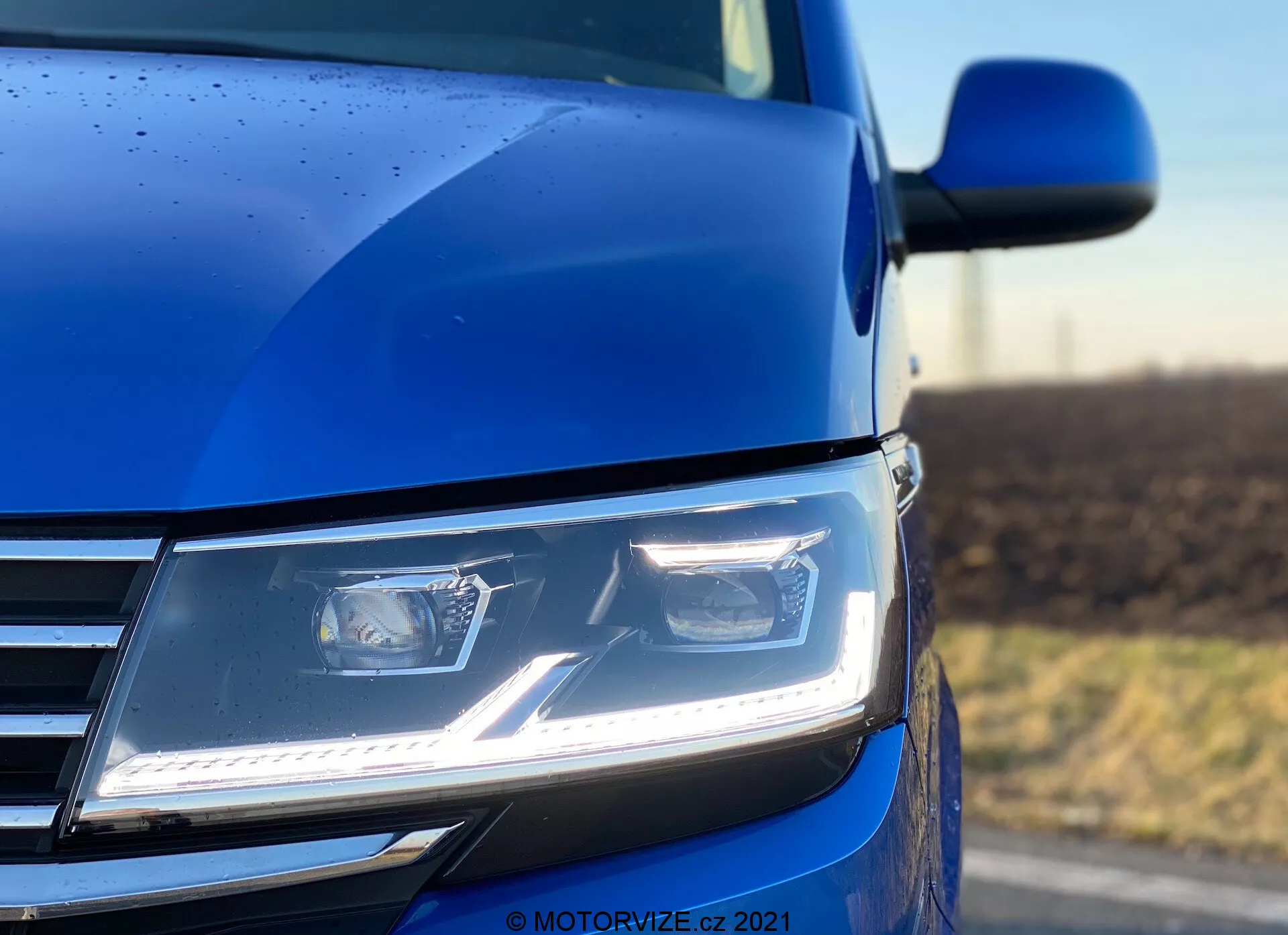 Una toma cercana y en ángulo bajo de la sección frontal izquierda del VW Nutzfahrzeuge Transporter 2019 (T6.1 Multivan) en azul, destacando los faros LED modernos y angulares con luces diurnas, parte de la parrilla cromada y un vistazo al contorno del capó y el espejo lateral. La foto, tomada con poca profundidad de campo, enfatiza el faro y la carrocería circundante, resaltando la tecnología avanzada de iluminación, el diseño contemporáneo y la calidad del acabado de la pintura, complementado por gotas de lluvia que indican una capa exterior detallada.