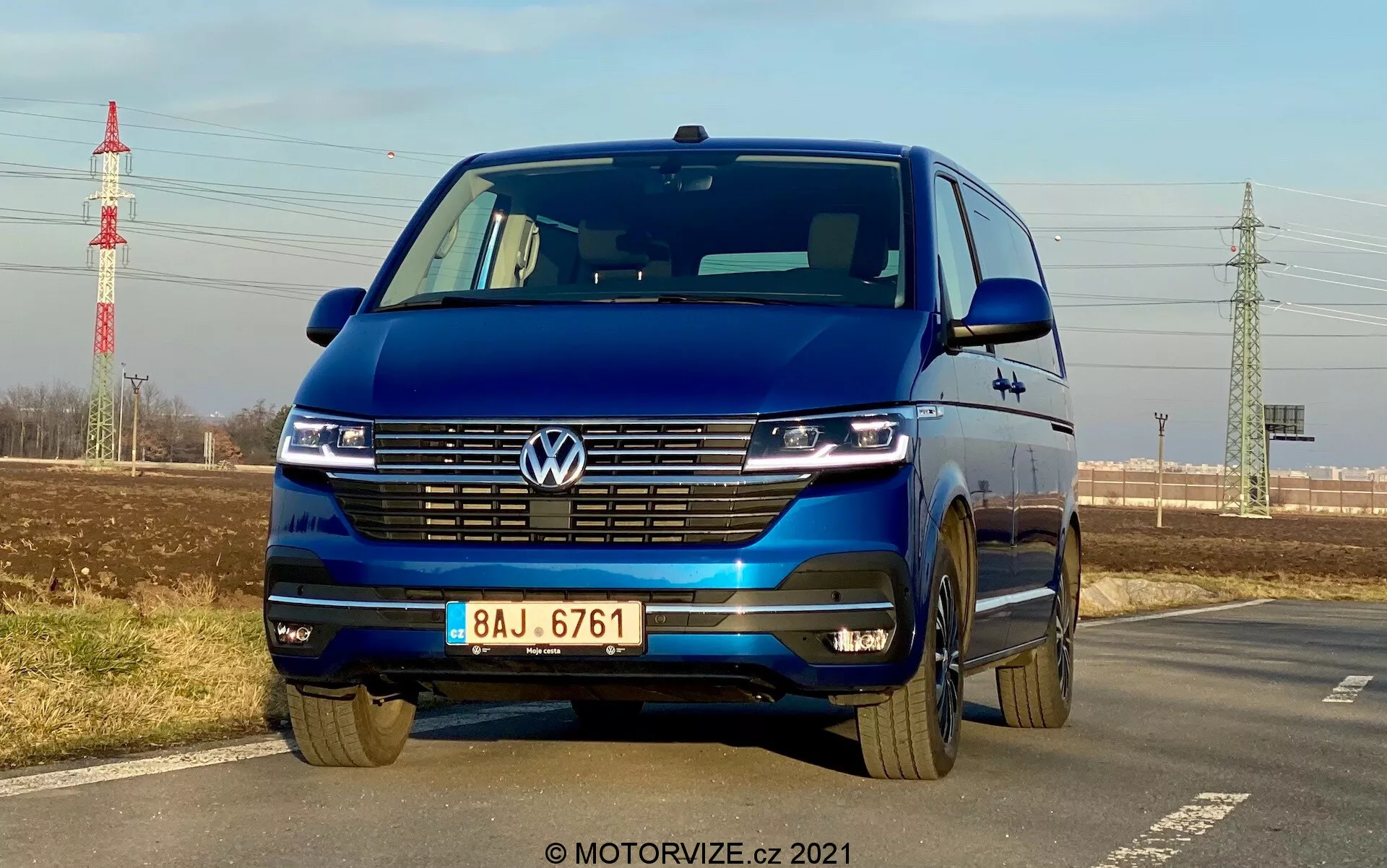 Volkswagen T6-1 Multivan 2019 azul capturado desde un ángulo frontal bajo destacando la parrilla, faros LED con luces diurnas, emblema de Volkswagen, parrilla ancha, parachoques bicolor y llantas de aleación; estacionado junto a una carretera con cielo claro, follaje distante, pilón eléctrico e infraestructura en el fondo.