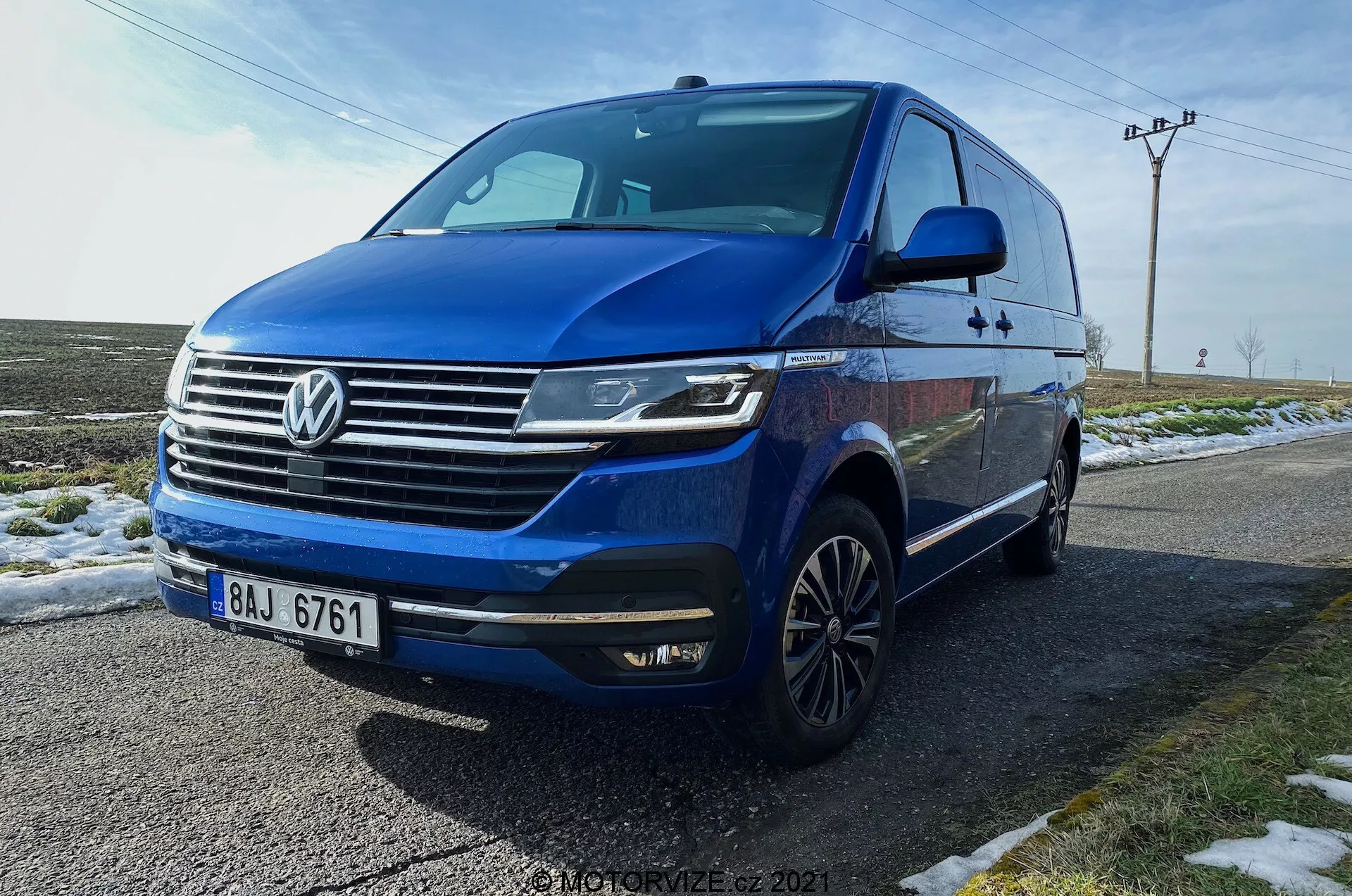 Volkswagen T6.1 Multivan Transporter 2019 azul capturado desde un ángulo bajo de tres cuartos delantero, mostrando la fascia frontal del auto, parrilla con el logo prominente de Volkswagen, barras horizontales cromadas, faros LED angulares con luces diurnas, parachoques delantero con luces antiniebla integradas y entradas de aire, línea dinámica que se extiende a lo largo de los lados, ruedas de aleación de varios radios, señales de giro integradas en los espejos laterales, ventanas tintadas. El vehículo está estacionado en una carretera mojada al lado de hierba nevada, con un fondo rural que incluye postes de servicios públicos y cielos despejados.