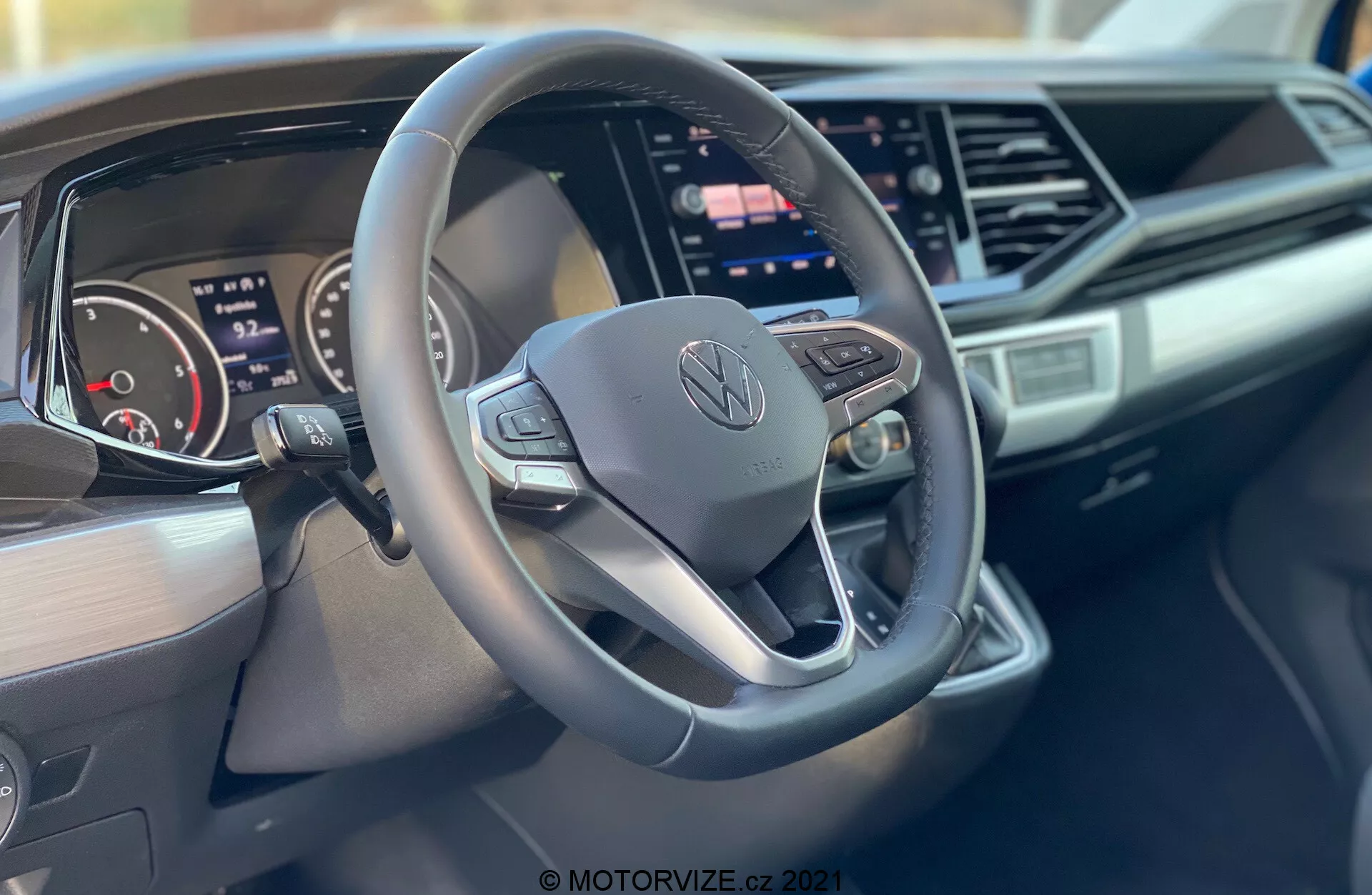 Imagen del interior de un Volkswagen Nutzfahrzeuge Transporter 2019 (T6-1 Multivan) centrándose en el volante y el tablero. El primer plano, tomado desde el lado del pasajero, destaca un volante de tres radios con fondo plano y botones de control con el logo central de Volkswagen, un cuadro de instrumentos digital, una gran pantalla táctil de infoentretenimiento, diseño elegante del tablero con acentos angulares y la marca en el lado del pasajero del tablero.