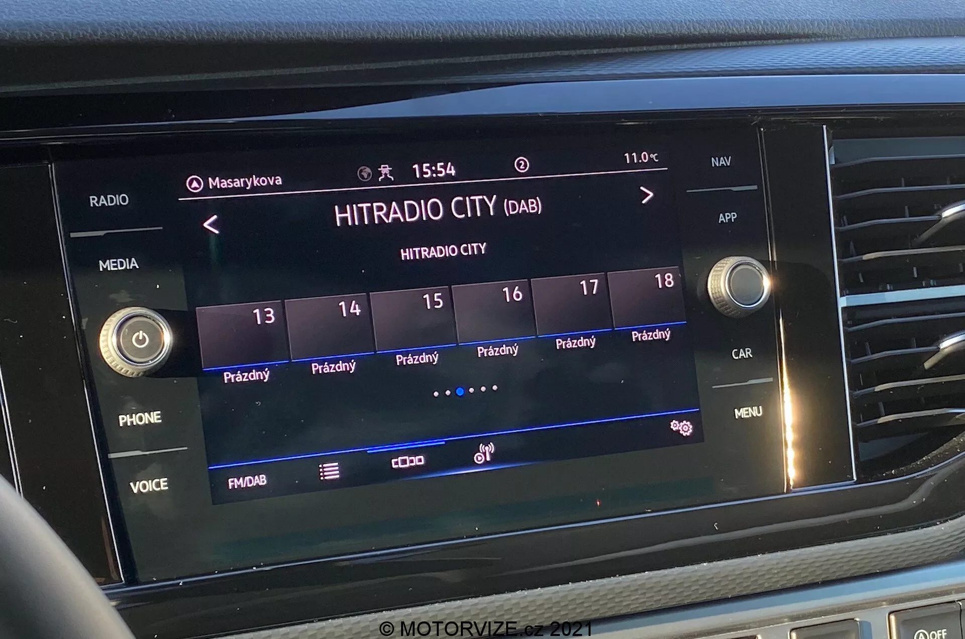 Vista frontal cercana de la pantalla del sistema de infoentretenimiento en el VW-Nutzfahrzeuge Transporter 2019 (T6-1 Multivan). La imagen muestra la estación de radio digital 'HITRADIO CITY' seleccionada, con varias opciones táctiles y botones de funciones como 'PHONE', 'VOICE' y 'MENU'. La pantalla muestra la hora '15:54' y la temperatura exterior '11.0°C', con iconos para perfiles de usuario, configuraciones del vehículo y fuerza de señal. La pantalla está integrada en la parte central del tablero, rodeada por botones táctiles y perillas de control.