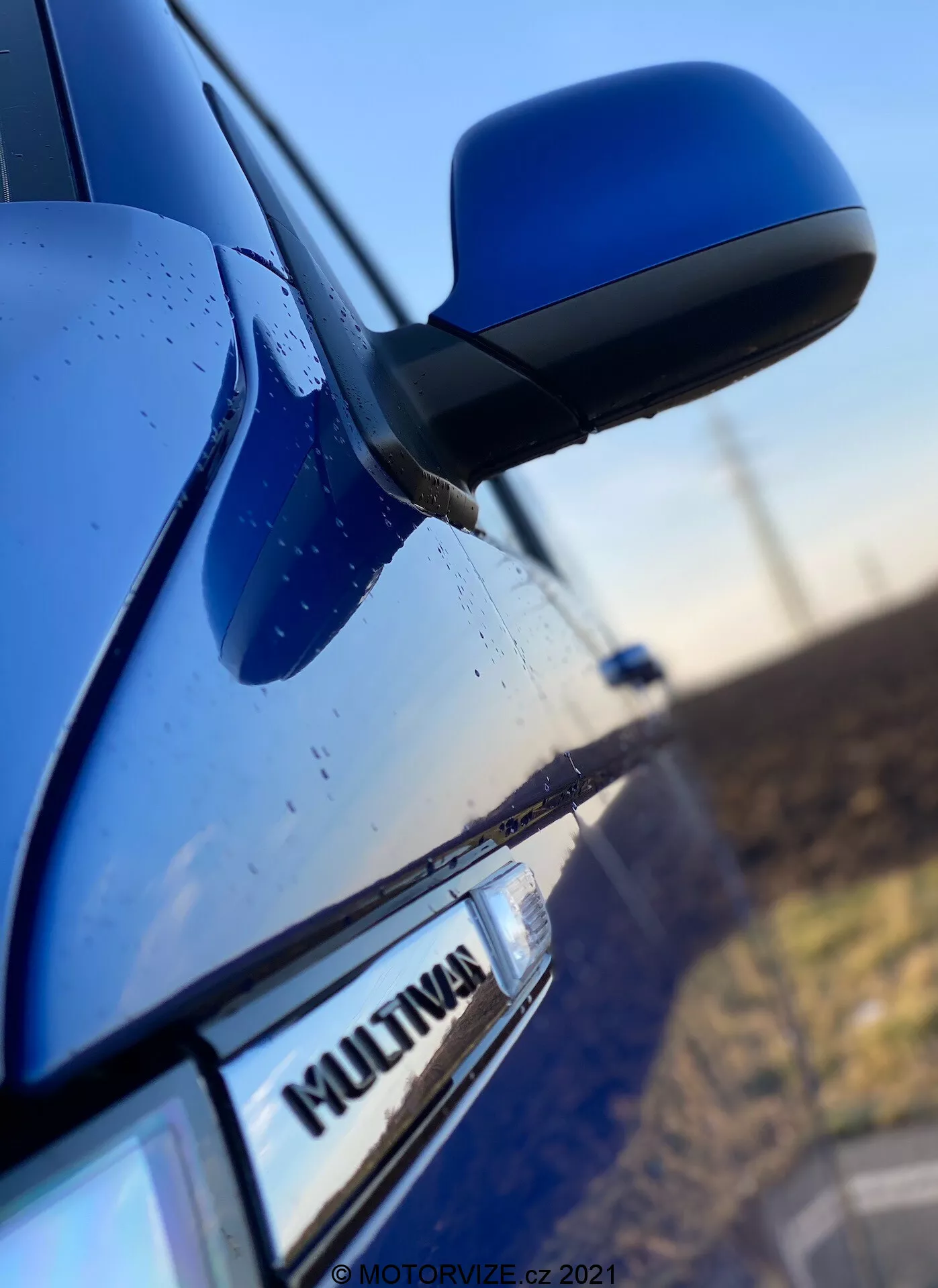 Fotografía en primer plano del lateral de un VW Nutzfahrzeuge Transporter 2019 azul (T6-1-Multivan) que muestra parte de la puerta del pasajero delantero, el espejo lateral y una parte de la puerta trasera. El foco está en la insignia 'MULTIVAN' del coche, con el espejo lateral aerodinámico ligeramente desenfocado. La carcasa del espejo coincide con el color del cuerpo del coche y las gotas de agua en la superficie sugieren características de impermeabilidad o lluvia reciente.