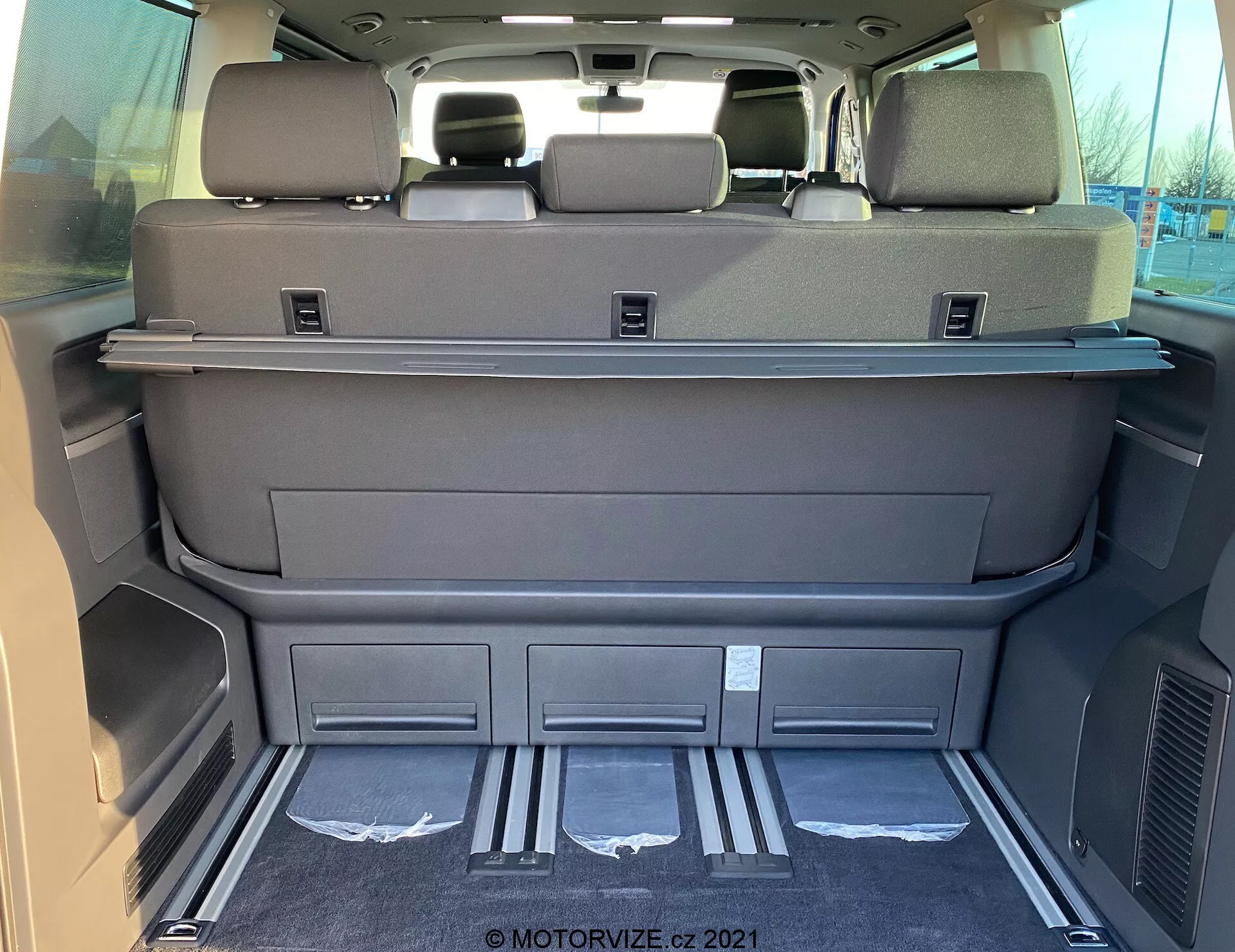Vista interior del espacio de carga de un VW Nutzfahrzeuge Transporter T6.1 Multivan 2019 desde la parte trasera, mostrando un área espaciosa con los asientos de la tercera fila abatidos, color gris oscuro o negro, estante cobertor de carga, reposacabezas en los asientos traseros, protectores metálicos de entrada, compartimentos moldeados, rejillas de ventilación, alfombrado limpio y posibles controles electrónicos de los asientos o portón trasero.