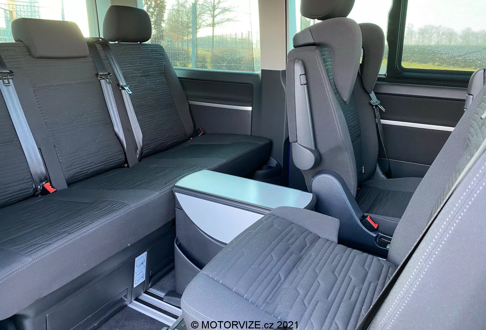Vista interior del VW-Nutzfahrzeuge Transporter 2019 (T6.1 Multivan) centrada en los asientos de la segunda y tercera fila. Fotografiado desde el lado a nivel de los ojos, la imagen muestra asientos individuales con reposacabezas prominentes en la segunda fila y un asiento de banco para tres pasajeros en la tercera fila. Los asientos son de tela bicolor y el piso presenta rieles metálicos para asientos reconfigurables. Se ven un reposabrazos plegable y hebillas de cinturones de seguridad. También se observa parcialmente la ventana y el almacenamiento lateral.