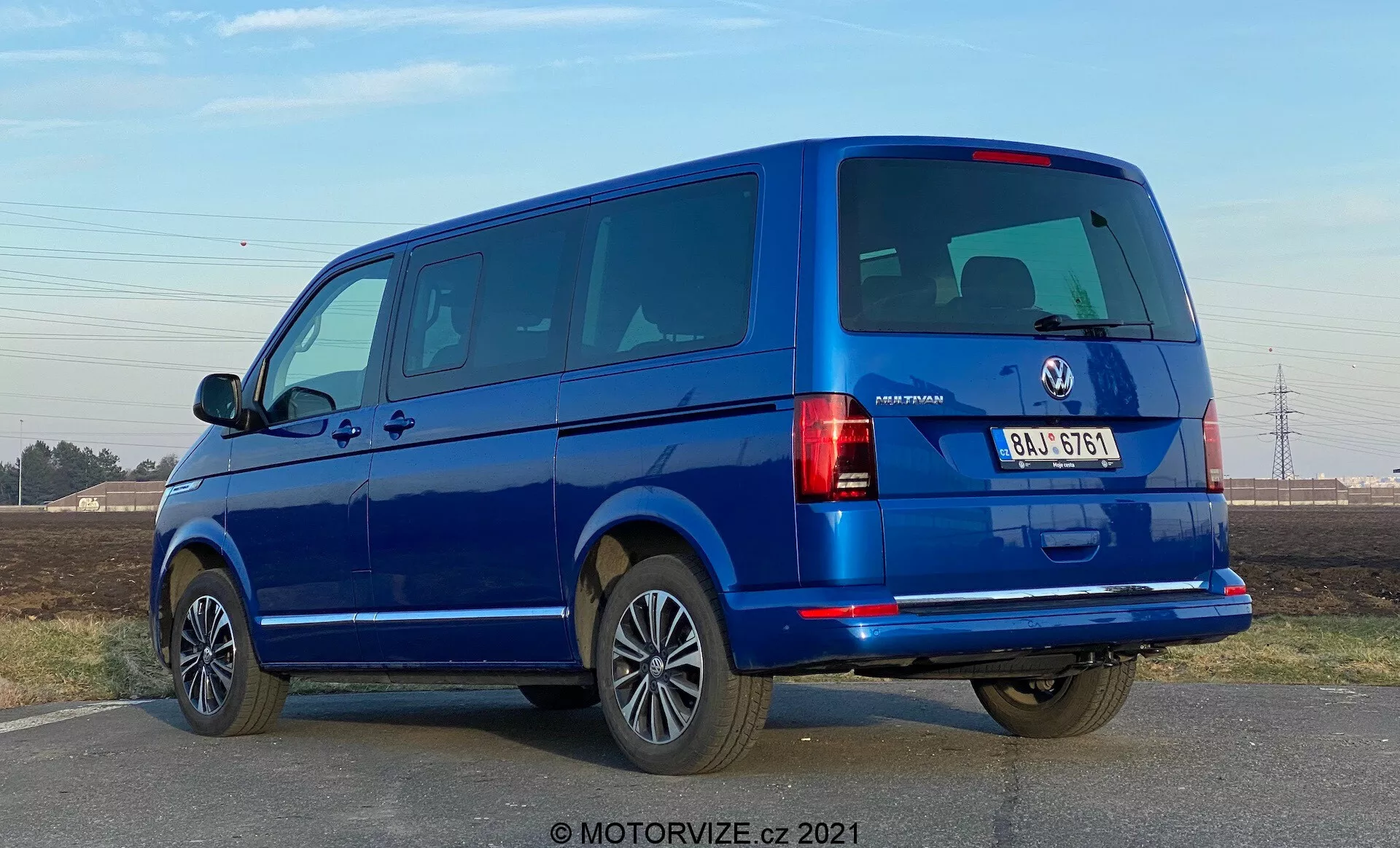 Una furgoneta azul VW-Nutzfahrzeuge Transporter 2019 (T6-1 Multivan) vista desde un ángulo trasero de tres cuartos. La imagen captura los grupos ópticos traseros, las ventanas traseras y la puerta trasera, así como el perfil lateral con puertas y ventanas de pasajero. La furgoneta ocupa una gran parte del marco a nivel de los ojos, revelando puertas traseras divididas y varias ventanas laterales, lo que indica un modelo para pasajeros. Las llantas de aleación, los parachoques del color de la carrocería y los espejos laterales sugieren un nivel de equipamiento alto. La iluminación sugiere que es de día con cielos despejados y hay sombras nítidas bajo el vehículo.