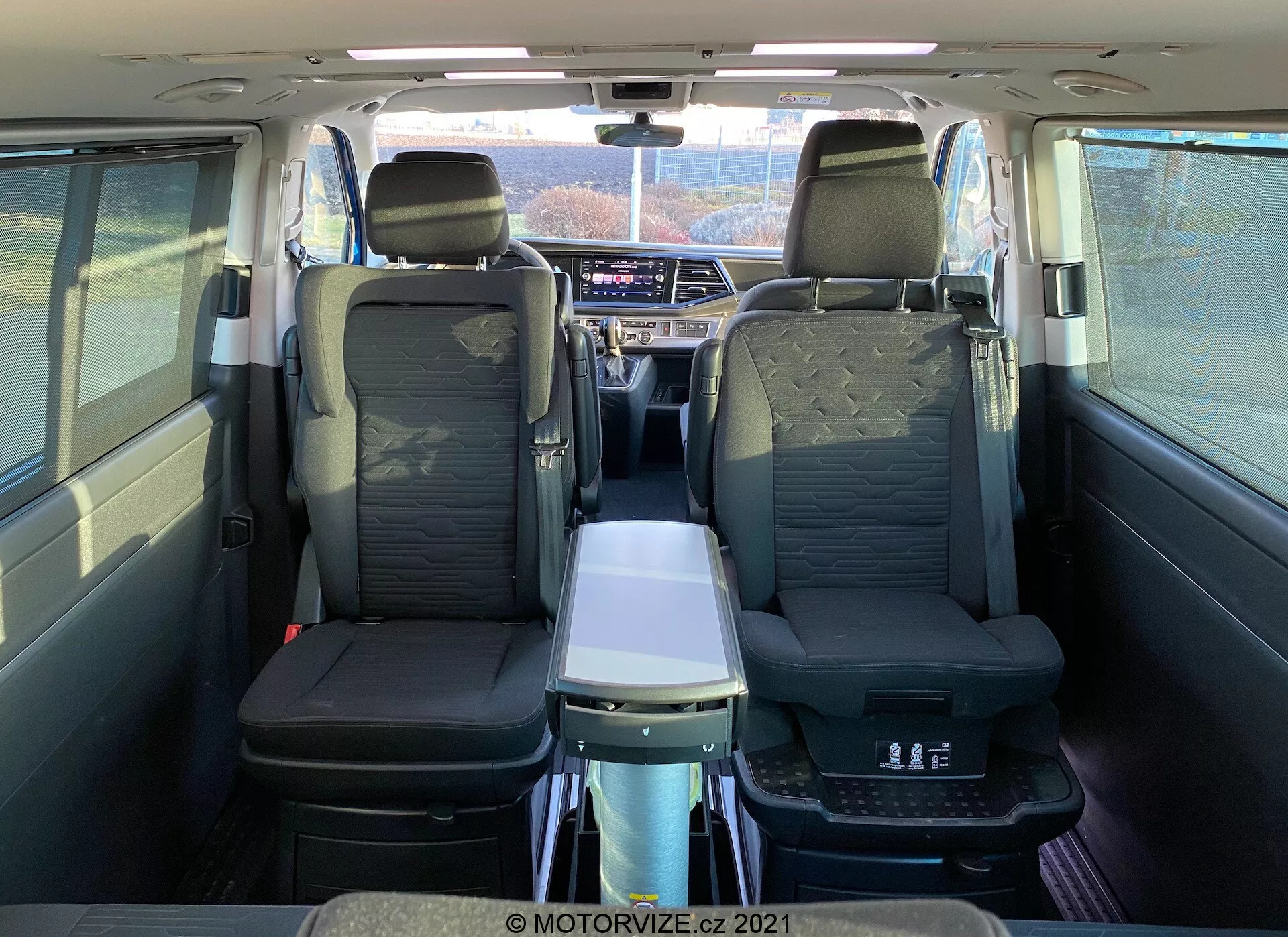 Interior del VW Nutzfahrzeuge Transporter T6-1 Multivan 2019 visto desde atrás, mostrando asientos de cuero, reposabrazos en la segunda fila, mesa plegable, techo panorámico, salidas de aire individuales y avanzado sistema de infoentretenimiento; destaca el confort de los pasajeros y el espacioso habitáculo.