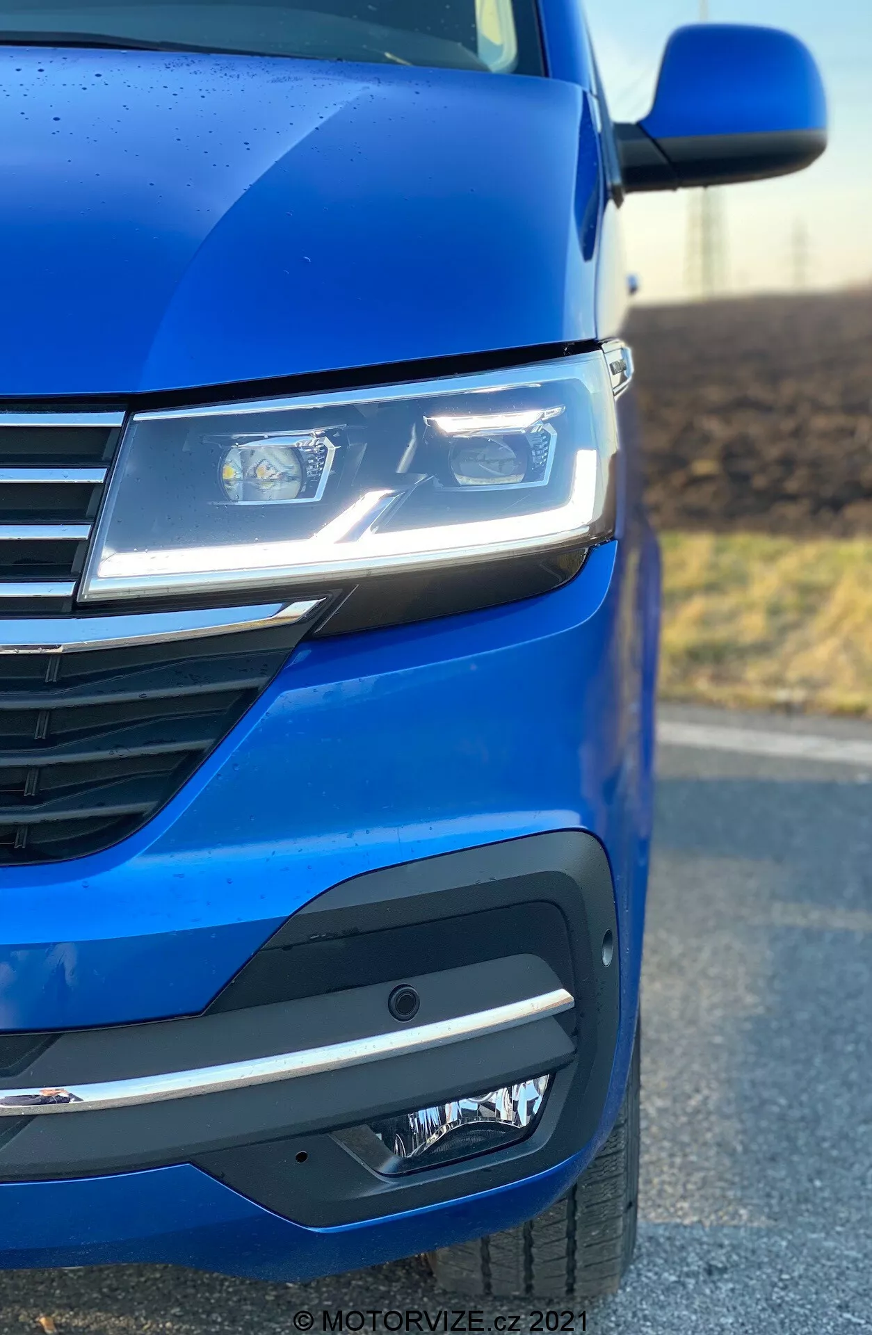 Primer plano del lado delantero izquierdo de un VW Nutzfahrzeuge Transporter 2019 (T6.1 Multivan) destacando la pintura azul metálico, los faros LED avanzados, el diseño moderno de la parrilla con líneas horizontales y el sofisticado parachoques delantero con luces antiniebla o unidades de sensor. El ángulo de la foto enfatiza la aerodinámica y los detalles de diseño del automóvil.