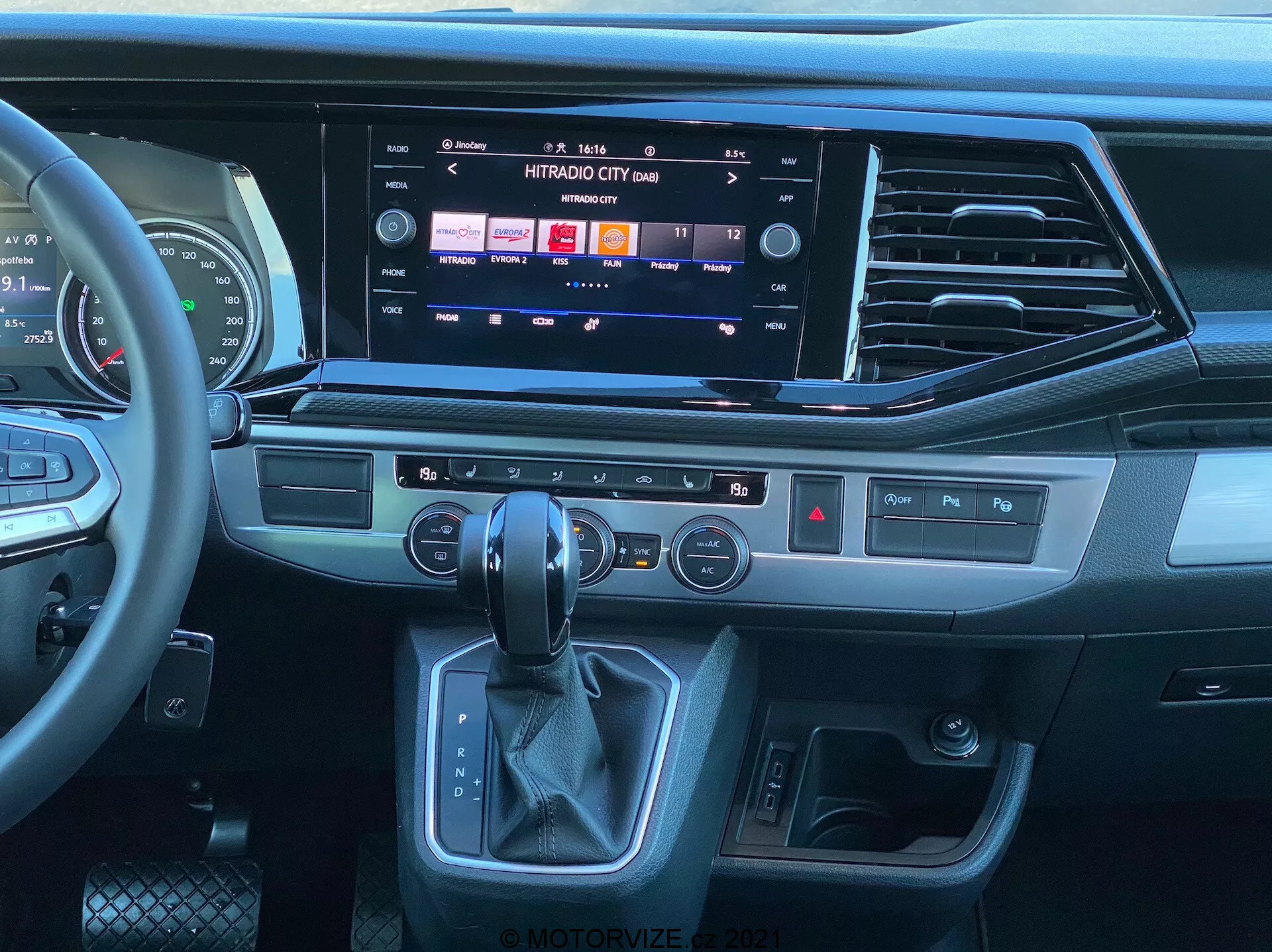 Vista interior del VW Nutzfahrzeuge Transporter 2019 T6.1 Multivan desde la perspectiva del asiento del pasajero, mostrando el moderno tablero y la consola central. Volante multifuncional cubierto de cuero con controles de medios, gran sistema de infoentretenimiento con pantalla táctil mostrando opciones de medios y conectividad, elegantes salidas de aire, botones táctiles para funciones rápidas, panel de control de clima de doble zona, área ergonómica de la palanca de cambios automática y botón de arranque/parada sin llave. El diseño presenta una mezcla de plásticos oscuros, adornos que parecen metal cepillado y materiales similares al cuero, destacando un ambiente contemporáneo y premium.