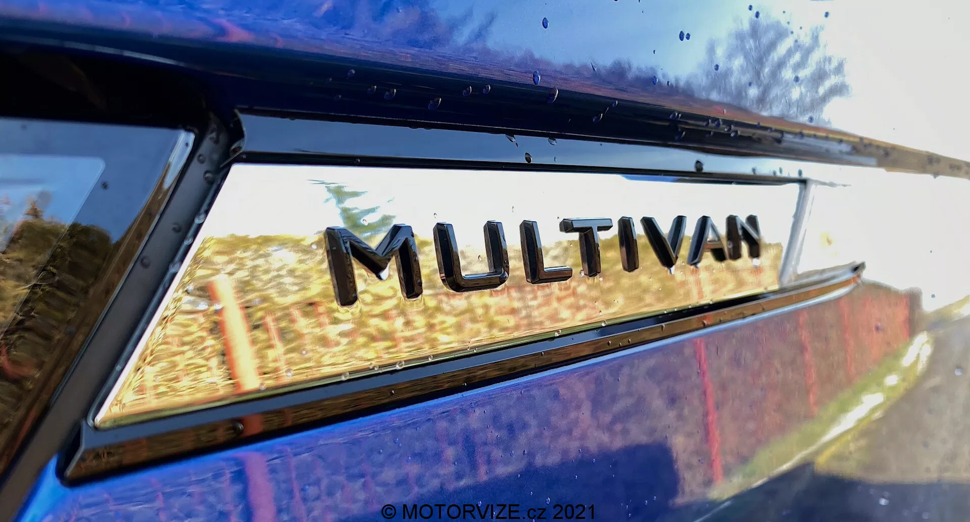 Vista de cerca de una parte del VW Nutzfahrzeuge Transporter 2019 (T6-1 Multivan) centrada en la insignia 'MULTIVAN'. La imagen está tomada desde un ángulo trasero bajo, mirando ligeramente hacia arriba donde la ventana trasera se encuentra con el borde del techo. Reflejos leves en el cristal de la ventana sugieren un entorno exterior. Esta imagen recortada de cerca resalta el emblema de designación del modelo del fabricante.