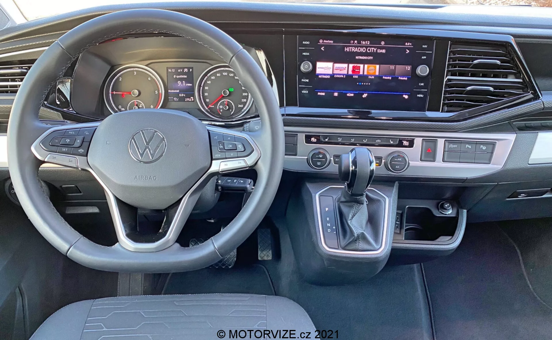 Vista interior del VW Nutzfahrzeuge Transporter 2019 T6.1 Multivan desde la perspectiva del asiento del conductor. Presenta un volante con la marca Volkswagen y botones multifuncionales, medidores analógicos, pantalla digital, consola central con sistema de infoentretenimiento con pantalla táctil activa, control de clima de doble zona, palanca de cambios con botón de estacionamiento electrónico y acentos modernos de metal cepillado.