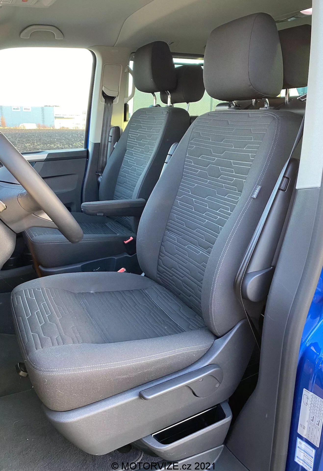 Interior del VW Nutzfahrzeuge Transporter 2019 T6.1 Multivan, mostrando el asiento delantero del pasajero y parte del asiento del conductor desde una perspectiva exterior del lado del pasajero. La toma, realizada desde un ángulo ligeramente bajo, resalta la tela texturizada con patrones geométricos en los asientos, la palanca de ajuste manual, los cinturones de seguridad y parte de la consola central y el volante. La luz natural ilumina el interior del vehículo.