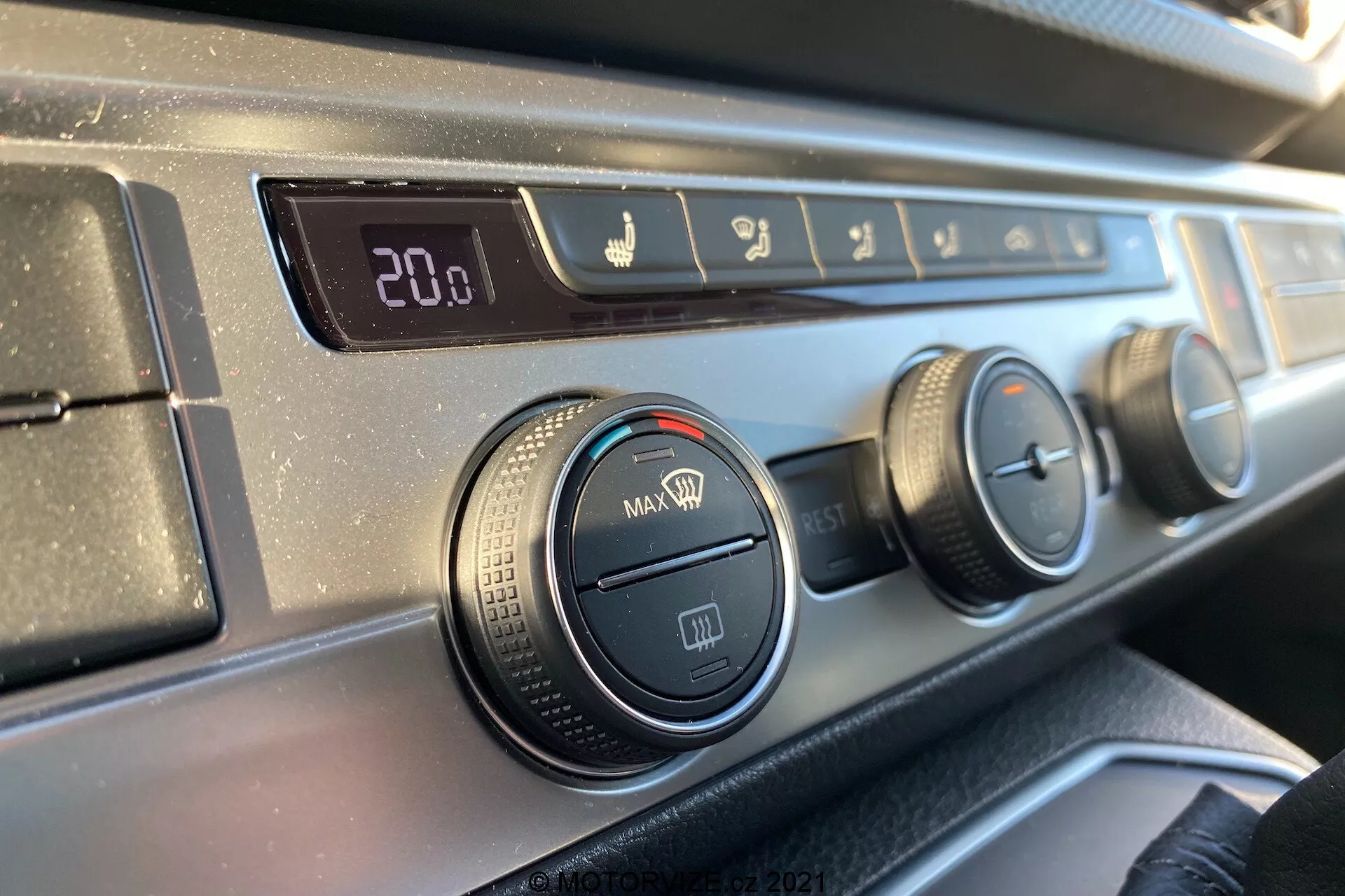 Vista en primer plano del panel de control del clima en el tablero del VW Transporter T6.1 Multivan 2019 desde la perspectiva de un ocupante del asiento delantero, con una pantalla de temperatura digital fijada en 20.0 grados Celsius, controles de dirección del flujo de aire, tres diales giratorios para ajustes de aire acondicionado y ventilador, y un botón de recirculación, todo con un borde de plástico texturizado negro y un acabado de tablero oscuro y liso.