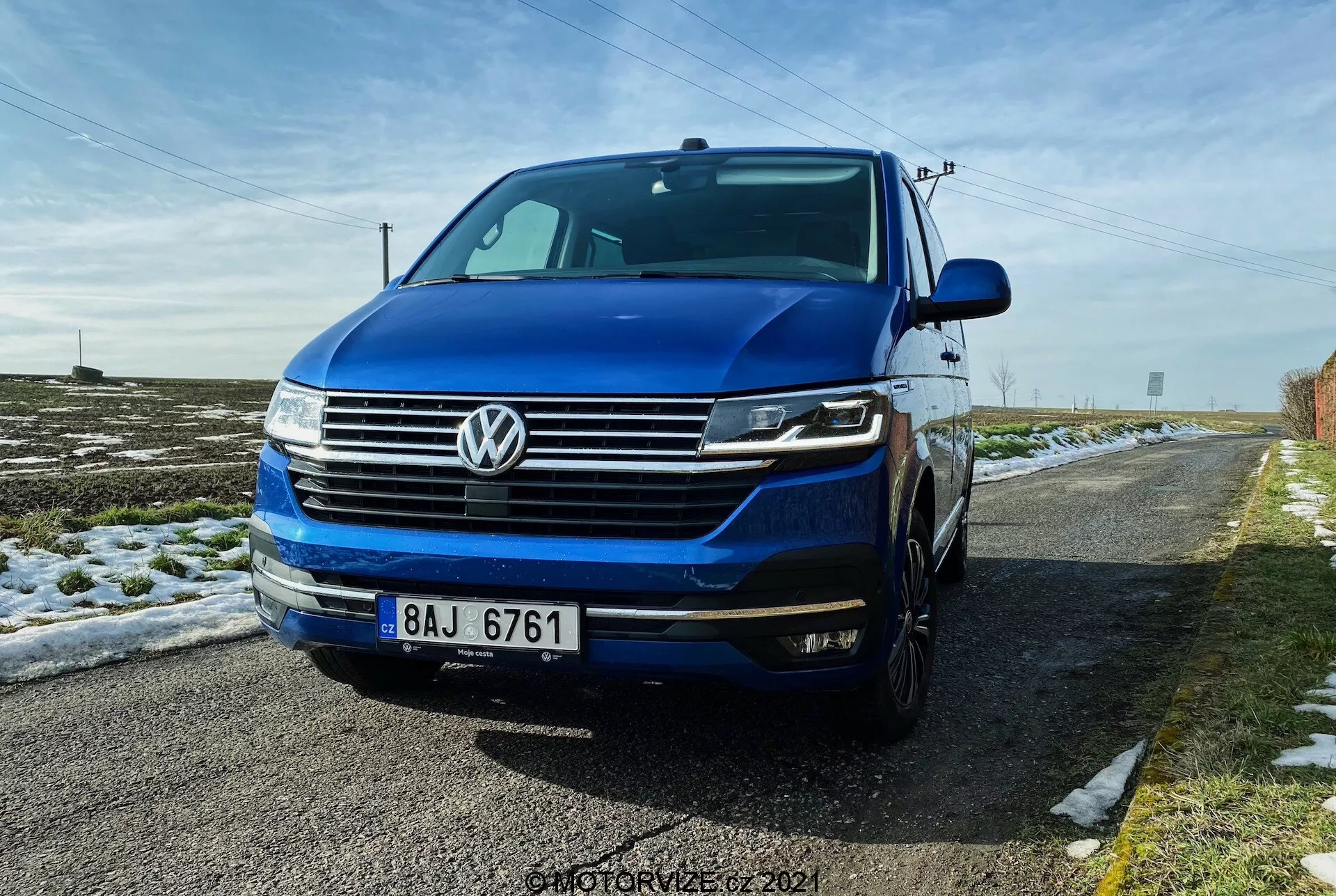 Vista tres cuartos frontal bajo del Volkswagen T6.1 Multivan 2019, con la parrilla delantera con líneas horizontales, el emblema distintivo de VW centrado en la parrilla, faros posiblemente con luces diurnas LED y luces bi-xenón o LED, y el parachoques delantero con luces antiniebla y entradas de aire. La imagen muestra parte del lado izquierdo, incluida la puerta del conductor y el guardabarros, con el capó y el parabrisas visibles. El coche es azul profundo y está estacionado en una carretera rural con nieve en el suelo y un cielo azul claro arriba.