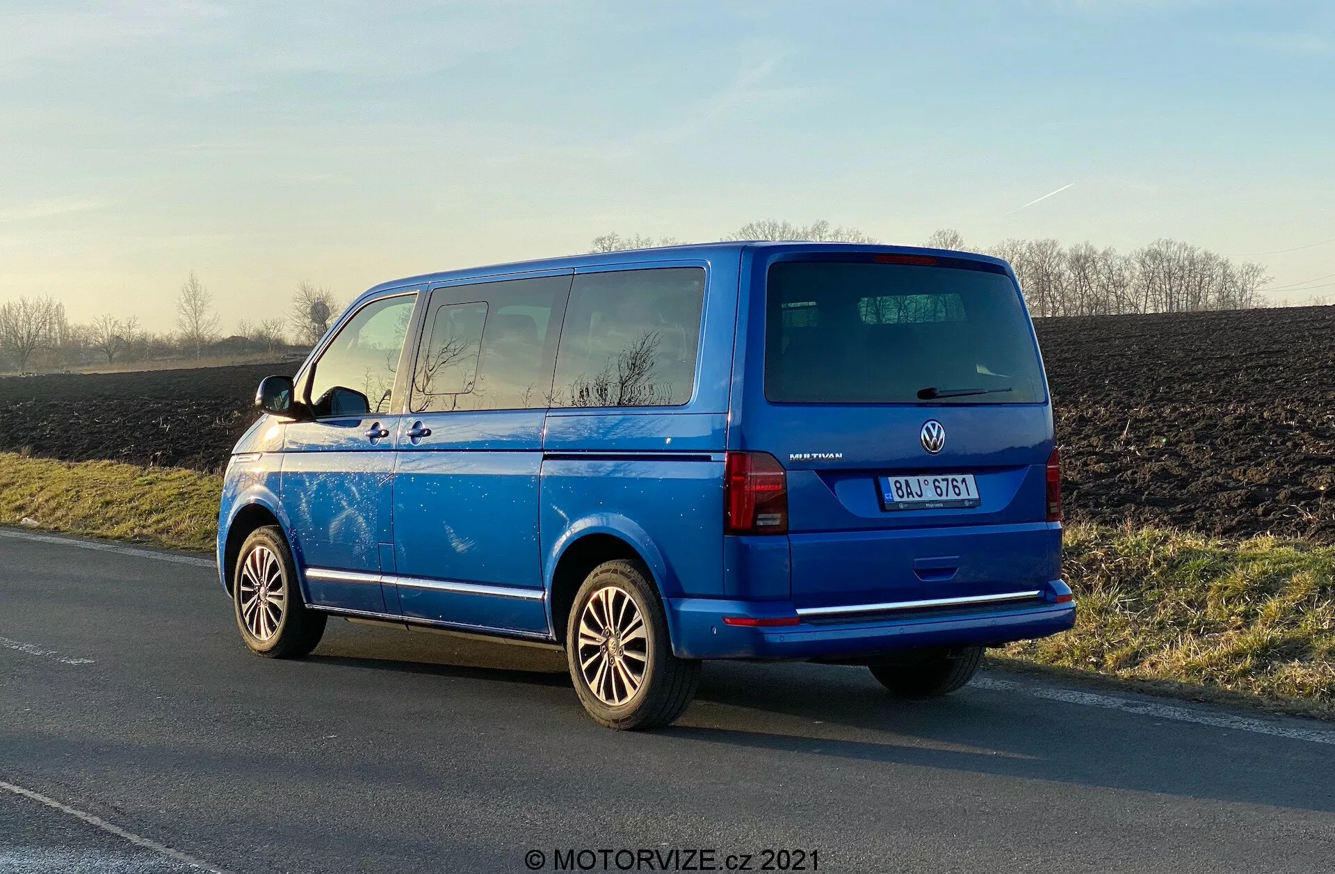 Volkswagen Transporter 2019 (T6-1 Multivan) de color azul vibrante estacionado en el borde de la carretera, vista trasera de tres cuartos mostrando las luces traseras, ventana trasera, puerta trasera, insignia del fabricante, detalles del modelo del vehículo; ruedas de aleación prominentes, espejos laterales externos, líneas desempañadoras de la ventana trasera, ventanas tintadas de la segunda fila, antena en el techo; estacionado a la luz del día con reflejos resaltando su superficie pulida, fondo rural.
