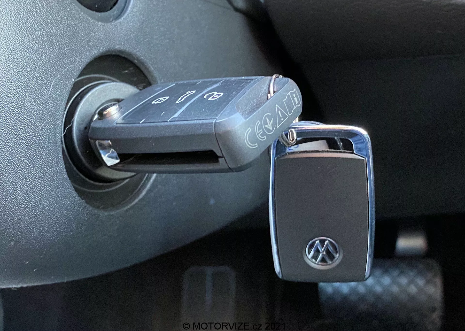 Primer plano de una llave plegable Volkswagen insertada en el interruptor de encendido de un VW Nutzfahrzeuge T6-1 Multivan de 2019. La llave, con botones para bloquear, desbloquear y alarma de pánico, muestra un logotipo de VW en el llavero. El interruptor de encendido está ubicado en el entorno de plástico oscuro de la columna de dirección, lo que indica que el encendido está en la posición 'apagado'. La foto, tomada desde un ángulo lateral con luz natural, resalta las texturas y acabados detallados del interior del vehículo.