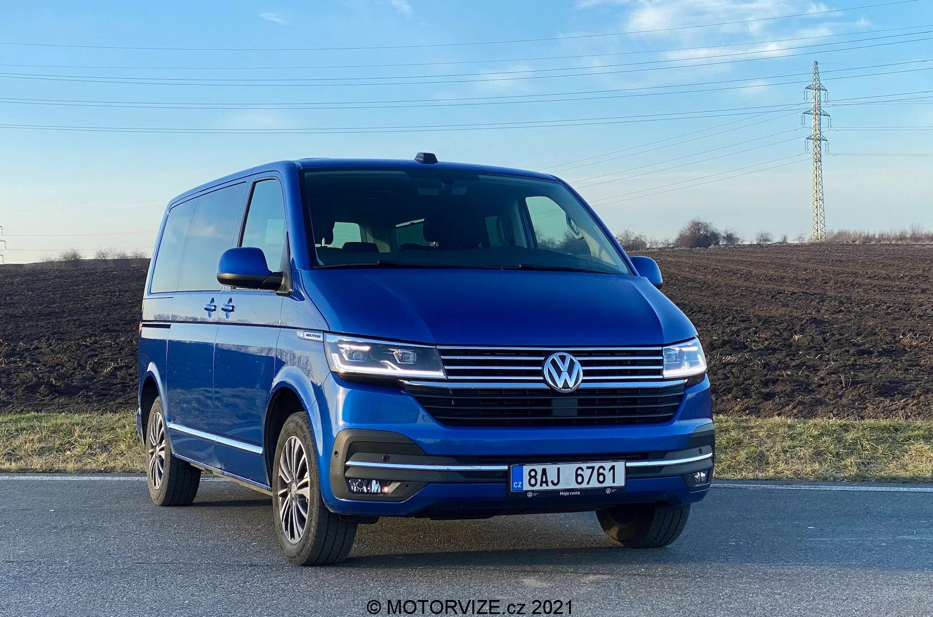 La imagen muestra una furgoneta azul Volkswagen VW-Nutzfahrzeuge Transporter 2019 (T6-1 Multivan) capturada desde un ángulo frontal bajo, destacando el cuarto frontal izquierdo con una vista clara de su fascia delantera y el perfil del lado del conductor. El vehículo está en una carretera, resaltando elementos como la insignia prominente de Volkswagen, faros elegantes, líneas esculpidas en el capó, luces antiniebla integradas en el parachoques y ruedas de aleación de múltiples radios. La fotografía, tomada a la luz del día, enfatiza el diseño exterior de la furgoneta y tecnologías de asistencia al conductor, como los sensores de aparcamiento.