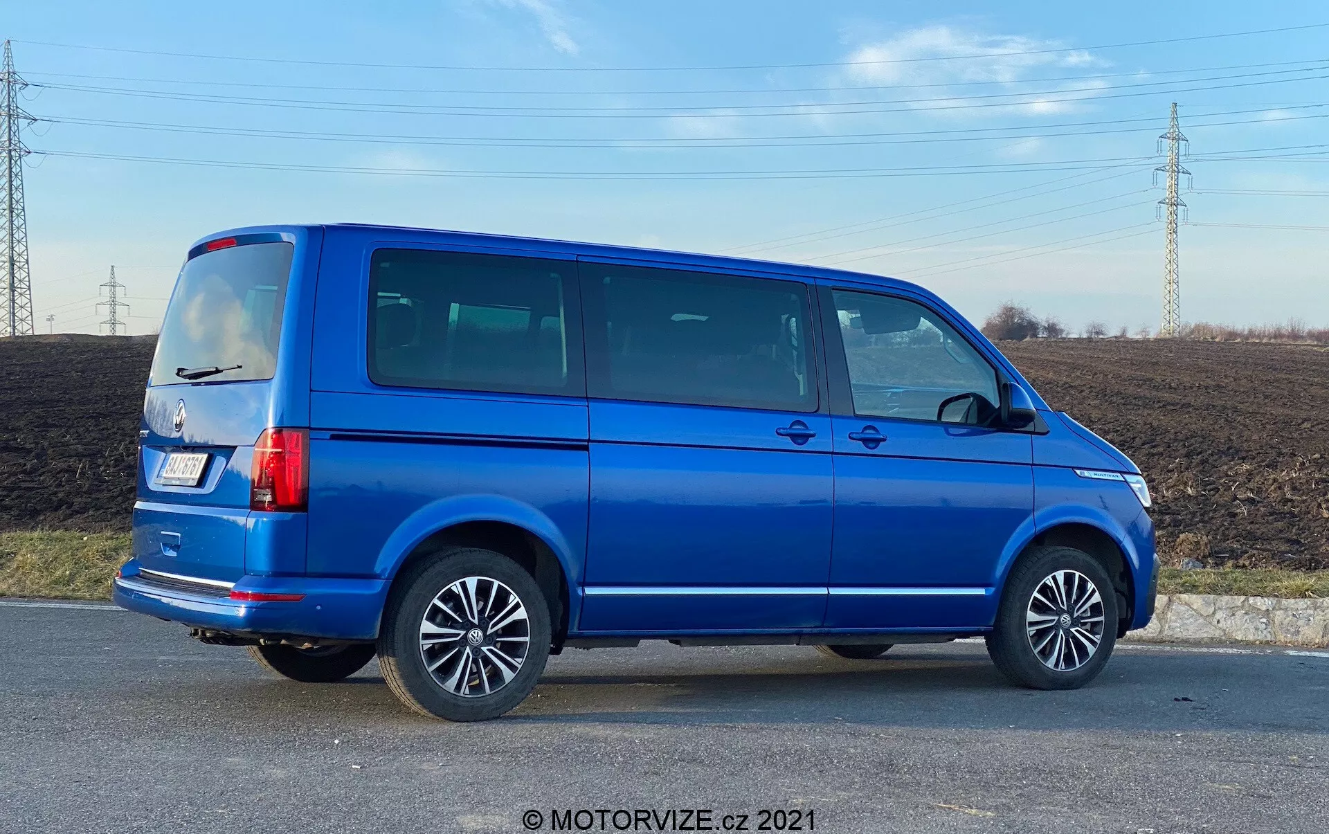 Vista completa de perfil lateral de un VW Nutzfahrzeuge Transporter azul de 2019 (T6.1 Multivan) con llantas de aleación, líneas visibles de puertas, ventanas laterales tintadas, silueta angular, luces traseras verticales, antena en el techo y pintura metálica azul brillante. La imagen está tomada en un ángulo que proporciona perspectiva tanto del lateral como de la parte trasera del vehículo, situada en una zona rural con un campo y líneas de alta tensión en el fondo, bajo luz diurna.
