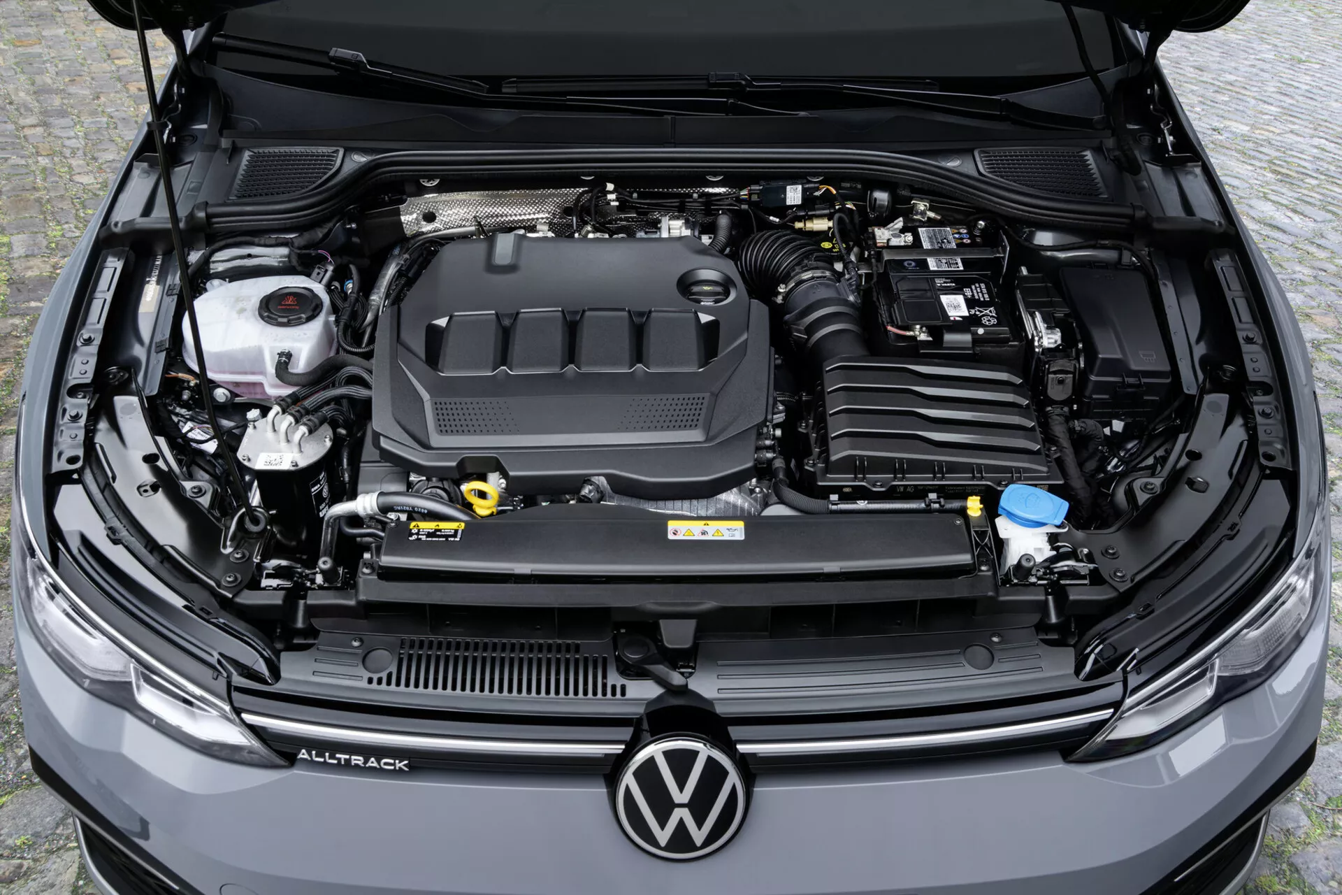 Vista aérea en ángulo alto del compartimiento del motor de un Volkswagen Golf Alltrack 2017 mostrando la cubierta del motor con logo, depósitos de líquidos y la batería del automóvil