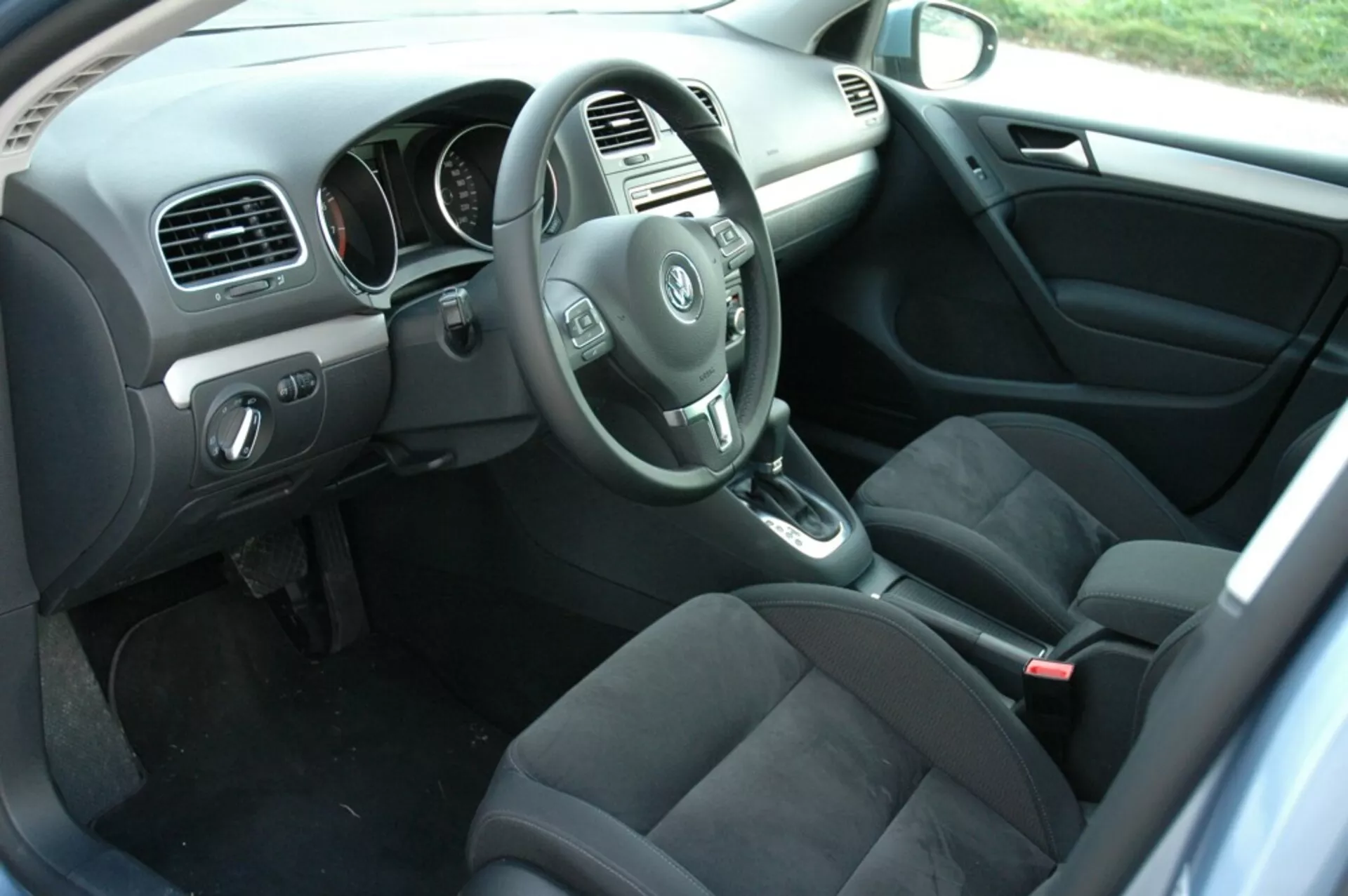 Interior del VW Golf 2008 mostrando el lado del conductor desde la perspectiva del pasajero trasero con el volante, tablero de instrumentos, consola central, palanca de cambios automática, asientos de tela oscura, controles en la puerta del conductor, suelo alfombrado con tapetes y luz natural