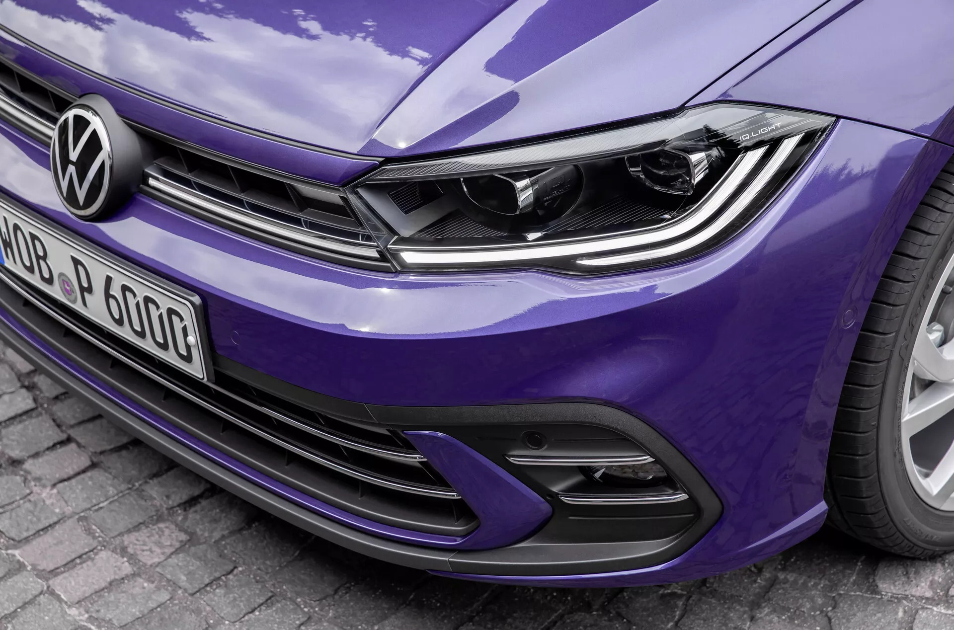 Vista frontal en primer plano, en ángulo, de un Volkswagen Polo 2021 VI-facelift azul, con un diseño moderno de faros con luces LED diurnas integradas, logotipo distintivo de Volkswagen en la parrilla de líneas horizontales y parachoques deportivo con tomas de aire y diseño de rejilla en forma de panal. Los detalles resaltan las líneas elegantes del capó y el acabado de pintura brillante, lo que sugiere una tecnología de iluminación avanzada y potencial asistencia al conductor.