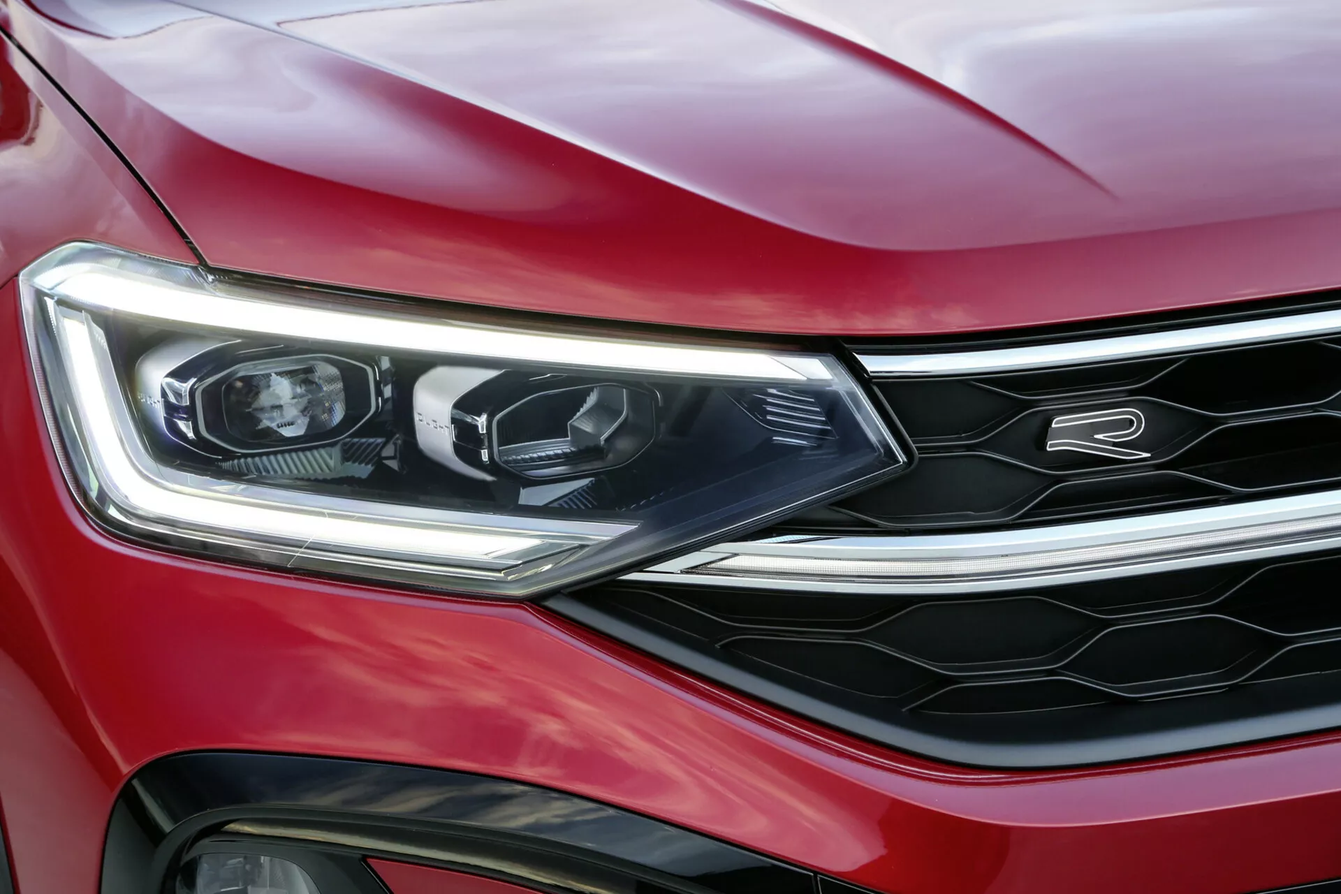 Primer plano de un VW Taigo 2021 en rojo, destacando el faro izquierdo, el parachoques delantero y una sección de la parrilla con acentos cromados, mostrando iluminación LED, diseño moderno y una apariencia deportiva y de lujo