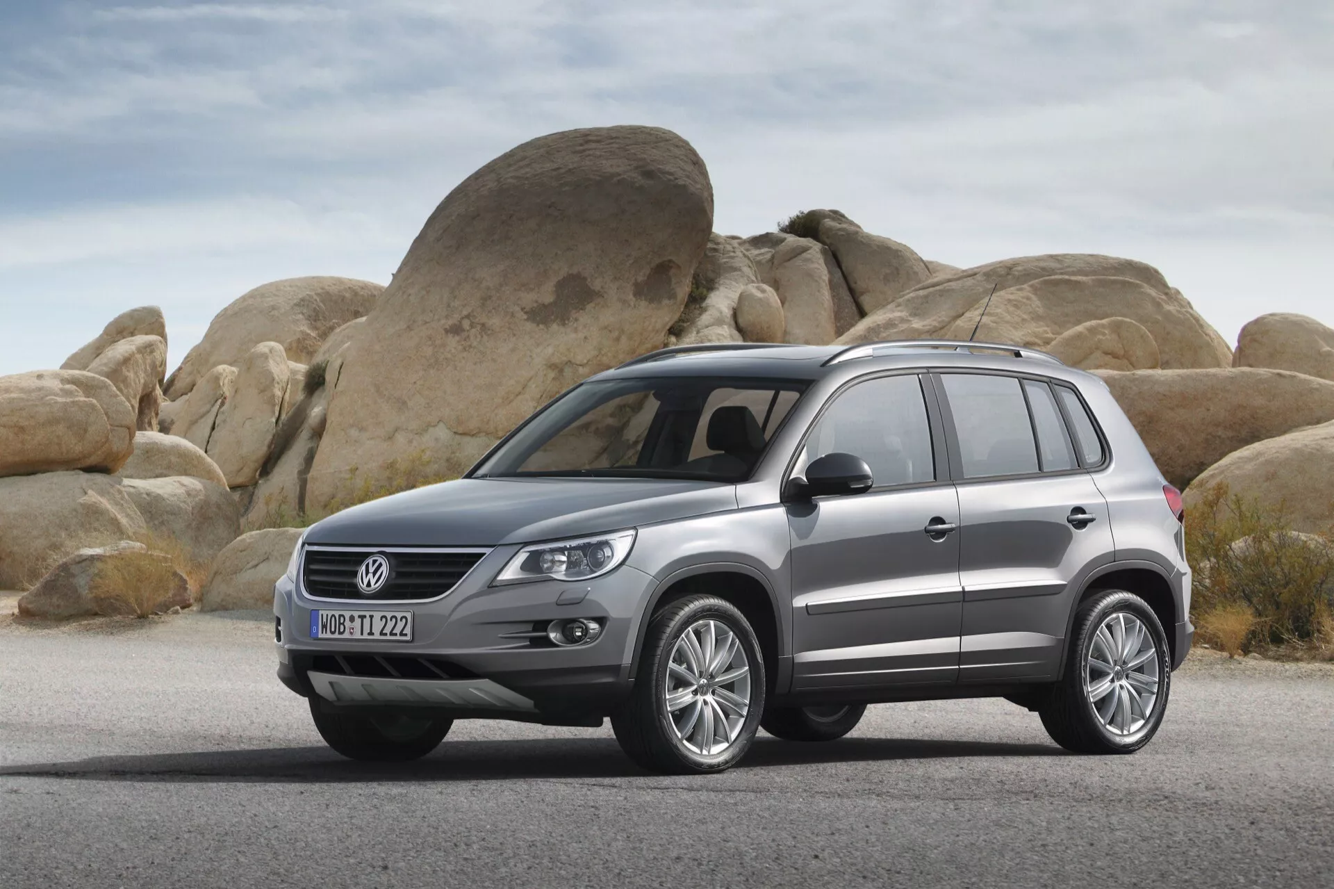 SUV Volkswagen Tiguan 2007 estacionado en una superficie seca y plana con grandes rocas redondeadas de fondo, mostrando el perfil frontal y lateral del vehículo incluyendo la parrilla, faros delanteros, parachoques delantero, capó, ruedas, paneles laterales, puertas y ventanas laterales con ruedas de aleación, barras de techo y acabado de pintura metálica.